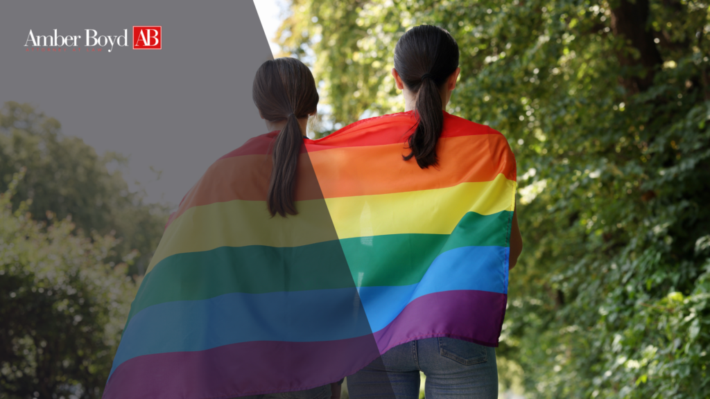 Two people seen from behind wrapped in a large rainbow Pride flag, outdoors, with the Amber Boyd AB logo in the top-left corner.