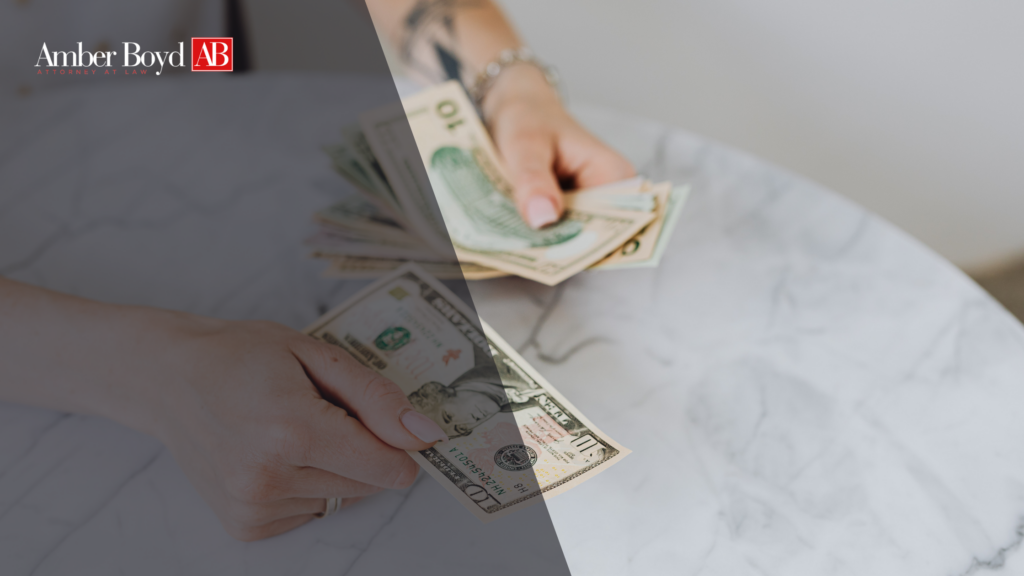 Hands counting several US dollar bills on a marble table, with Amber Boyd AB logo in the upper left corner (law firm image).