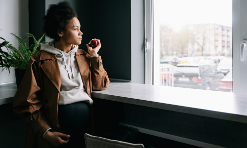 Woman seated by a cafe window, holding a red apple near her lips as she gazes outside.