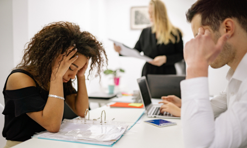 Frustrated woman at a desk with hands on her head; another stressed man sits nearby during a tense office meeting in the background.
