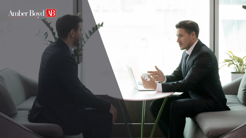 Two business professionals in suits discuss a topic at a glass table in a modern office, with a laptop between them and a logo visible in the corner.