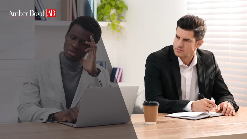 Two men sit at a desk in an office: one with a stressed expression looking at a laptop, the other in a suit taking notes.