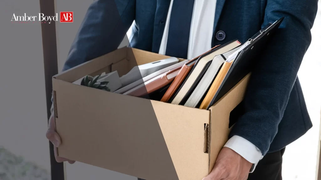 Person in a dark suit carries a cardboard box filled with folders and notebooks; Amber Boyd logo appears in the top-left corner.