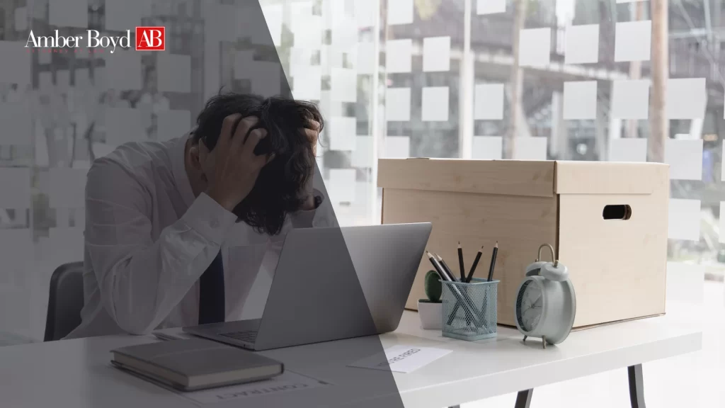 Overworked man at a desk, head in hands, laptop open, cluttered with office supplies and a large cardboard box in a glass-walled workspace.