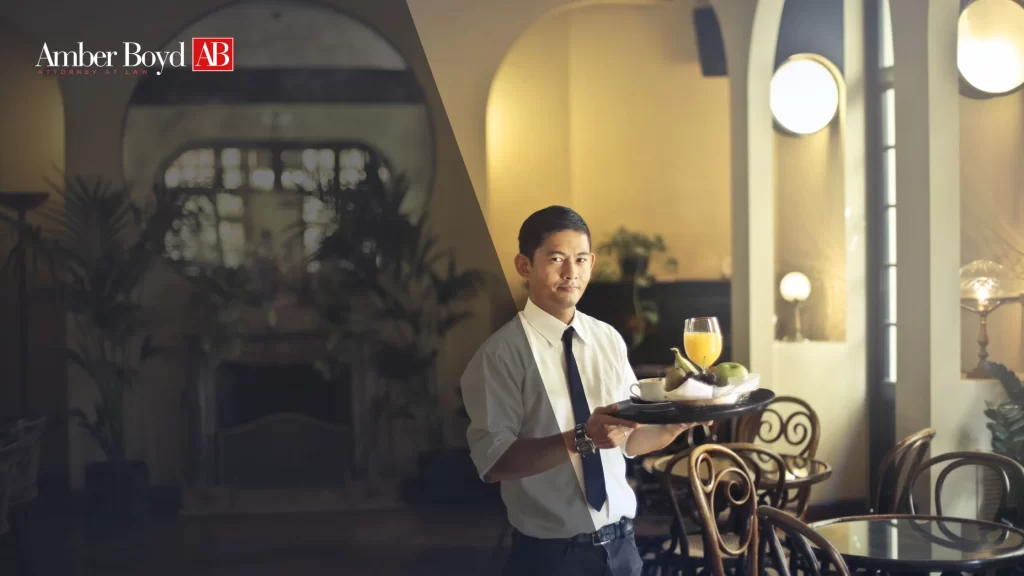 Indiana Tipped Employee Laws What Restaurants Owe You - Amber Boy - Amber Boy Waiter in a white shirt and black tie offers a breakfast tray with orange juice and fruit in a cafe, Amber Boyd branding visible on the left wall area.