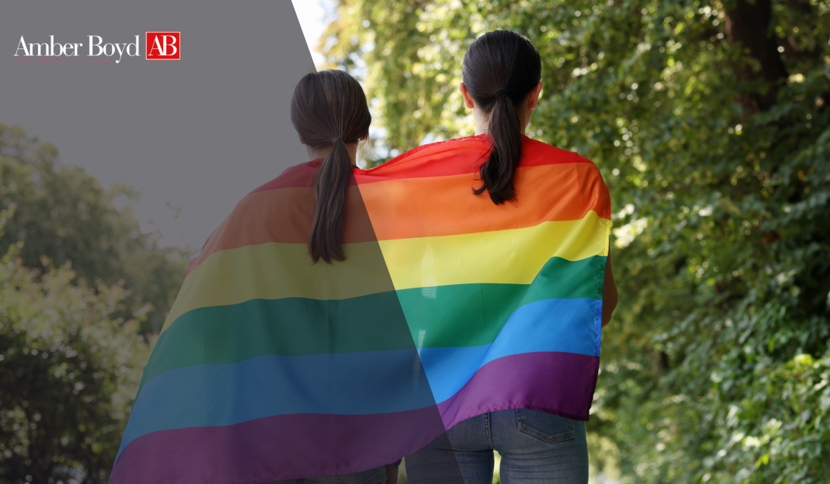 Two people seen from behind wrapped in a large rainbow Pride flag, outdoors, with the Amber Boyd AB logo in the top-left corner.