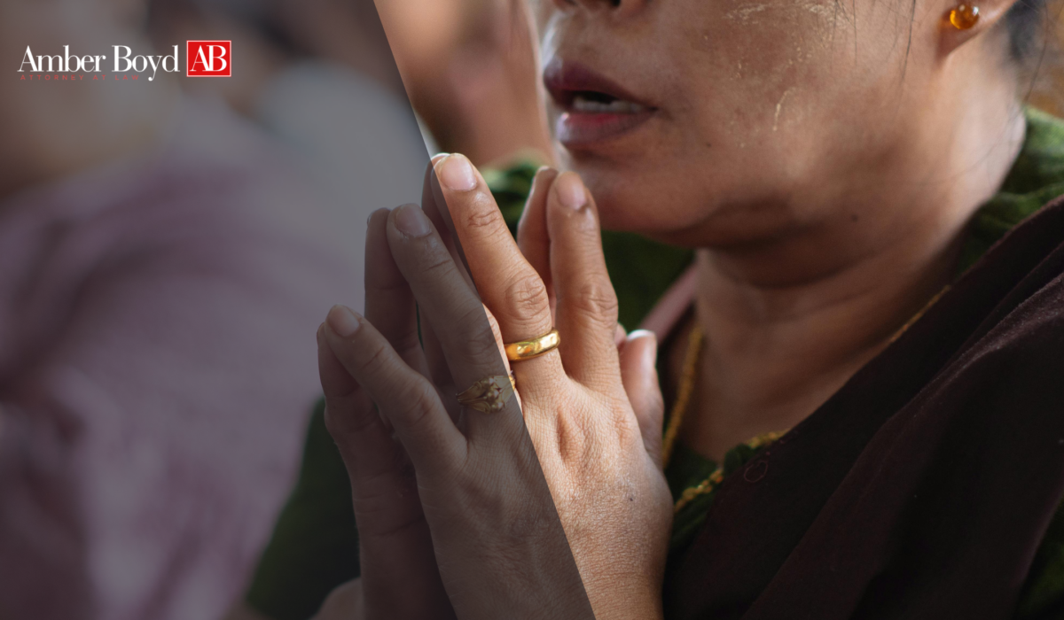 Close-up of hands pressed together in prayer with gold rings, partial face of a woman in background; Amber Boyd logo appears top-left.