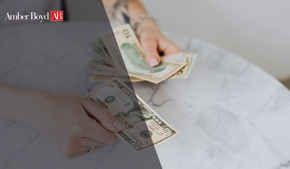 Hands counting several US dollar bills on a marble table, with Amber Boyd AB logo in the upper left corner (law firm image).