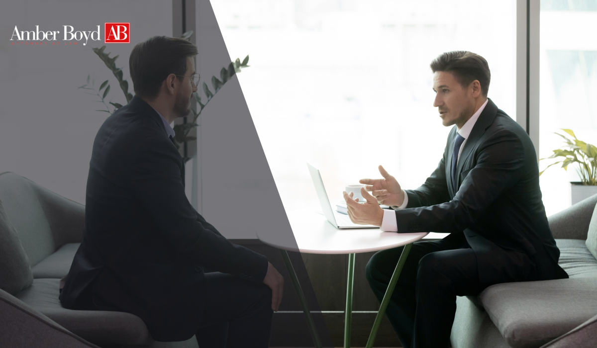 Two business professionals in suits discuss a topic at a glass table in a modern office, with a laptop between them and a logo visible in the corner.