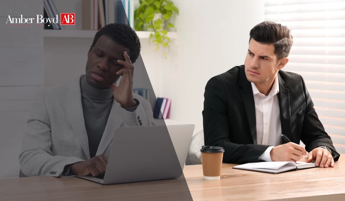 Two men sit at a desk in an office: one with a stressed expression looking at a laptop, the other in a suit taking notes.