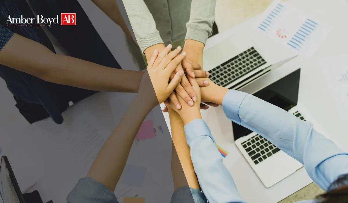 Team members stack hands in the center of a meeting table with laptops, showing collaboration; Amber Boyd logo in the corner.