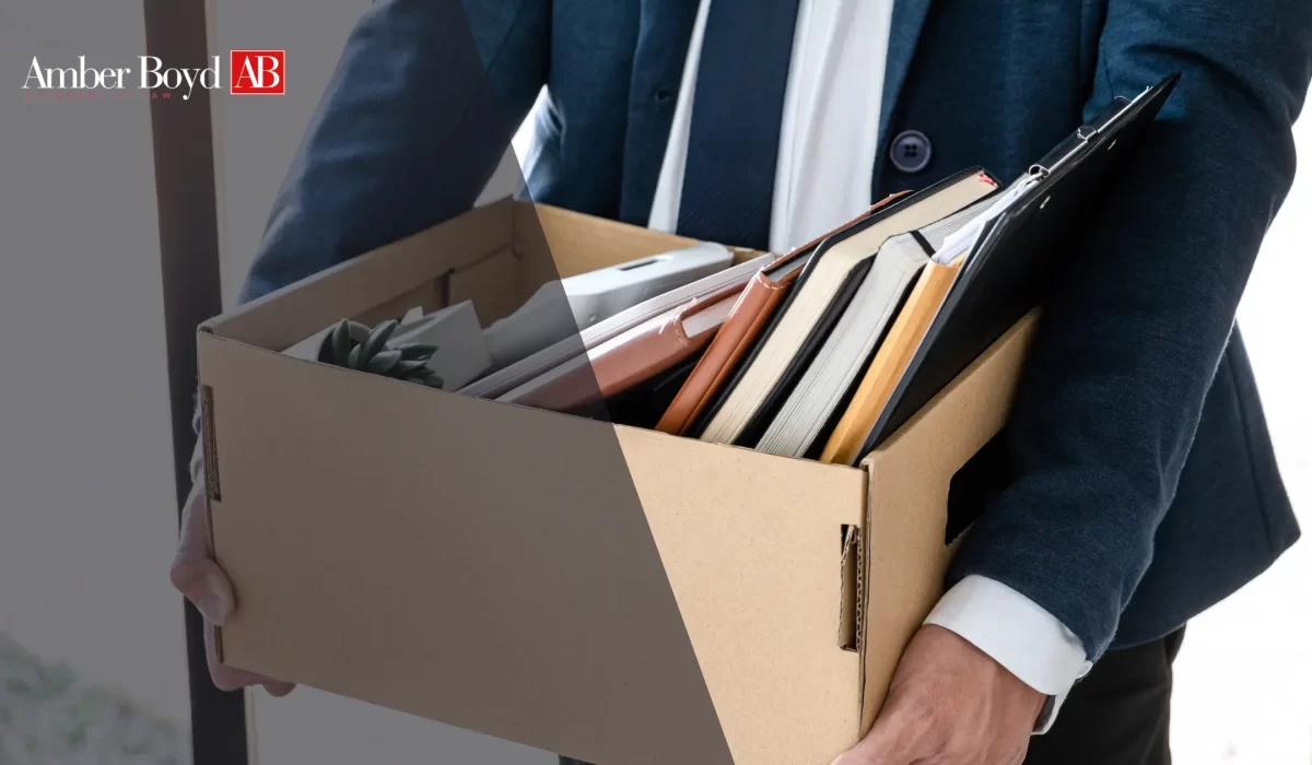 Person in a dark suit carries a cardboard box filled with folders and notebooks; Amber Boyd logo appears in the top-left corner.