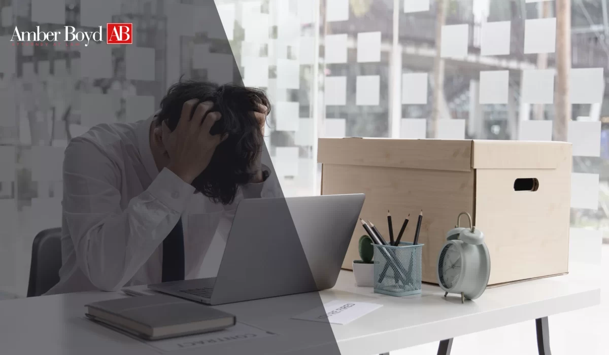 Overworked man at a desk, head in hands, laptop open, cluttered with office supplies and a large cardboard box in a glass-walled workspace.