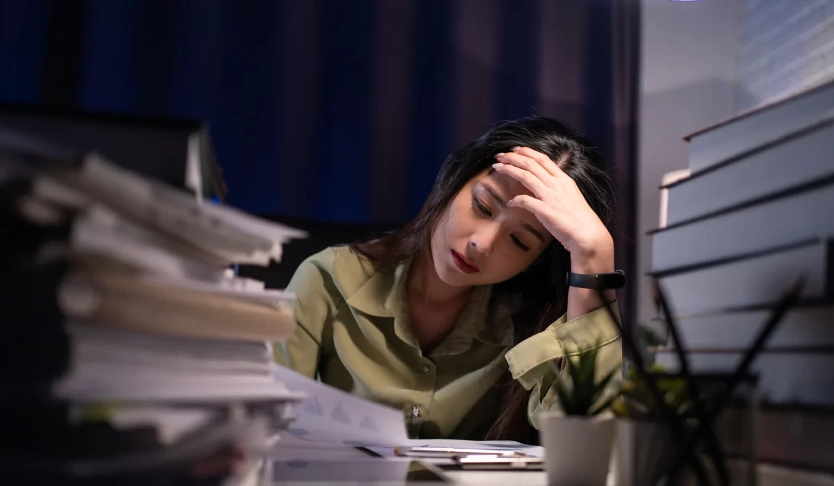 Woman with hand on forehead at a cluttered desk, surrounded by piles of papers, looking stressed or tired.