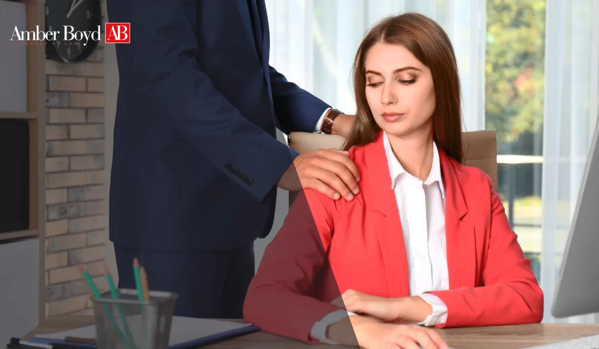 Woman in a red blazer sits at a desk while a man in a navy suit rests his hand on her shoulder; Amber Boyd AB logo in the top-left corner.