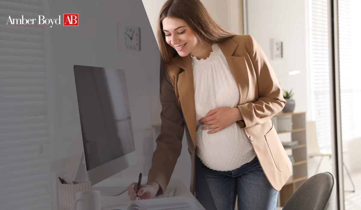Pregnant woman standing at a desk, writing with a pen beside a computer in a bright, modern office; Amber Boyd AB logo visible in the top-left corner.