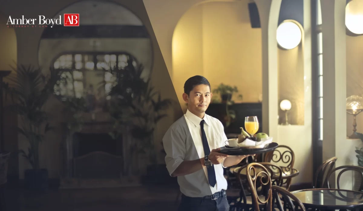 Waiter in a white shirt and black tie offers a breakfast tray with orange juice and fruit in a cafe, Amber Boyd branding visible on the left wall area.